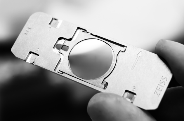 Hand holding metal ZEISS sample holder with central circular cutout and arrow marking
